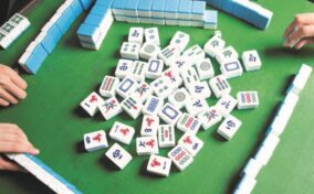 Three people play a game of mahjong, with tiles scattered on a green table and players' hands partially visible arranging their tiles.