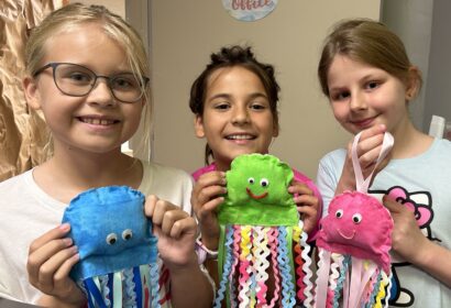 Three children smile while holding handmade jellyfish crafts with colorful ribbons and googly eyes.
