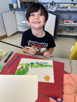 A young child sits at a table in a classroom, smiling while drawing a colorful outdoor scene with markers and colored pencils.