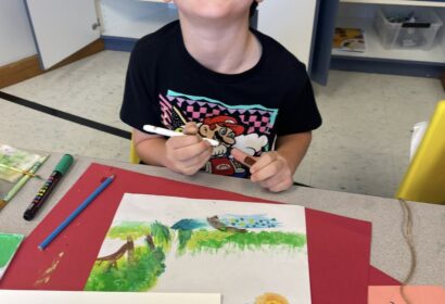 A young child sits at a table in a classroom, smiling while drawing a colorful outdoor scene with markers and colored pencils.