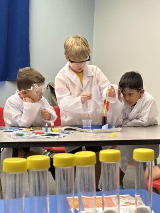Three children wearing lab coats and goggles conduct a science experiment at a table, using pipettes and test tubes, with more test tubes in the foreground.
