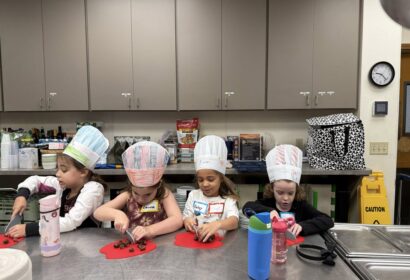 Four young children wearing paper chef hats sit at a kitchen counter, preparing food on red cutting boards with plastic knives.
