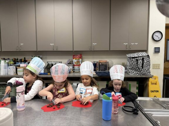 Four young children wearing paper chef hats sit at a kitchen counter, preparing food on red cutting boards with plastic knives.
