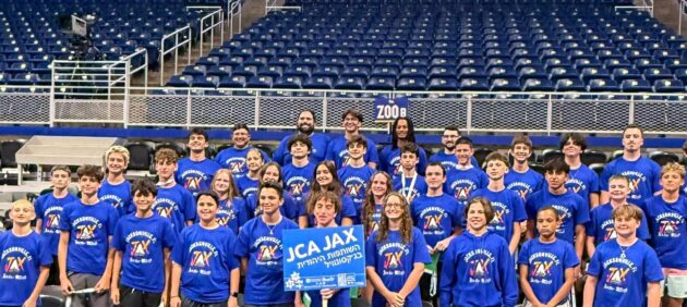A large group of people in matching blue shirts pose for a photo in an indoor arena, holding a "JCA JAX" sign in the front row.