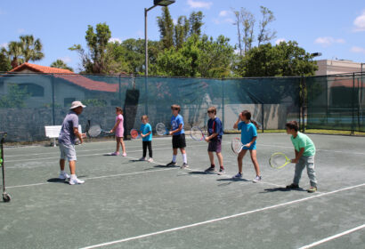 A tennis coach instructs six children lined up on an outdoor tennis court, each holding a racket and ready to play.