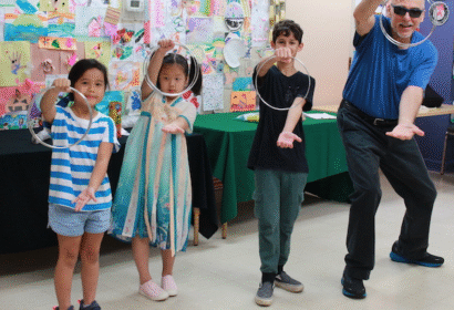 Three children and an adult pose indoors, each holding a metal ring in one hand, with colorful artwork displayed on the wall behind them.
