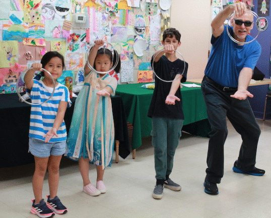Three children and an adult pose indoors, each holding a metal ring in one hand, with colorful artwork displayed on the wall behind them.