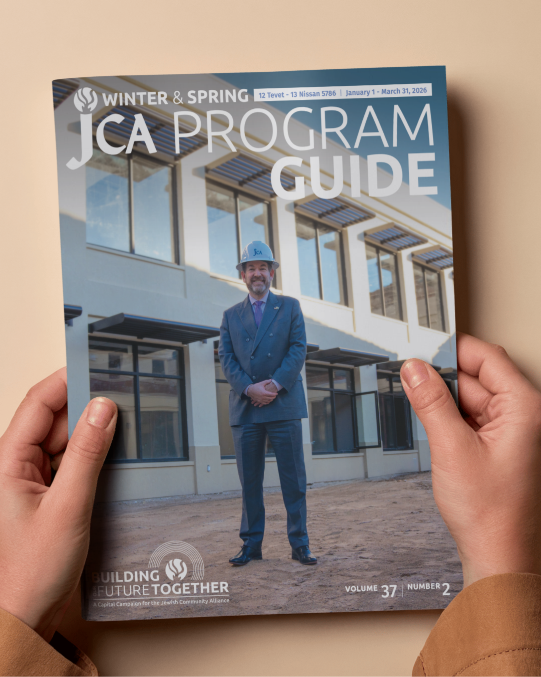 Person holding a magazine titled "JCA Program Guide" featuring a man in a suit and hard hat standing in front of a building; Winter & Spring 2026 edition.