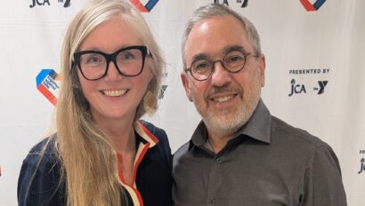 A woman and a man stand together smiling in front of a step-and-repeat banner with logos and hearts containing American flag elements.