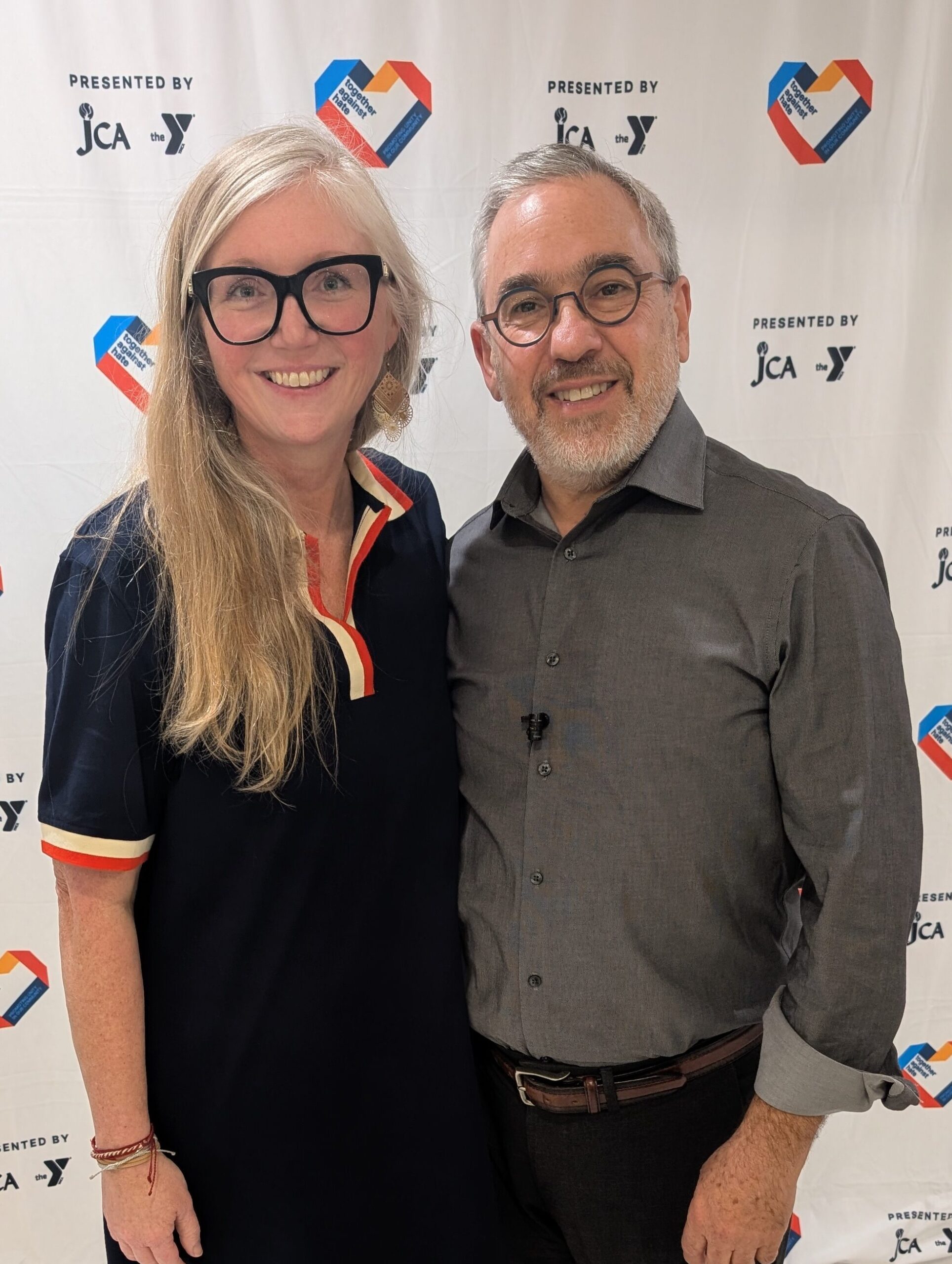 A woman and a man stand together smiling in front of a step-and-repeat banner with logos and hearts containing American flag elements.