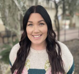 Woman with long dark hair, wearing a light sweater with a colorful geometric design, smiles at the camera outdoors with trees and blurred buildings in the background.