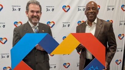 Two men in suits stand on a carpeted floor, holding a large, colorful heart-shaped sign that reads "Promoting Unity In Our Community" in front of a branded backdrop.