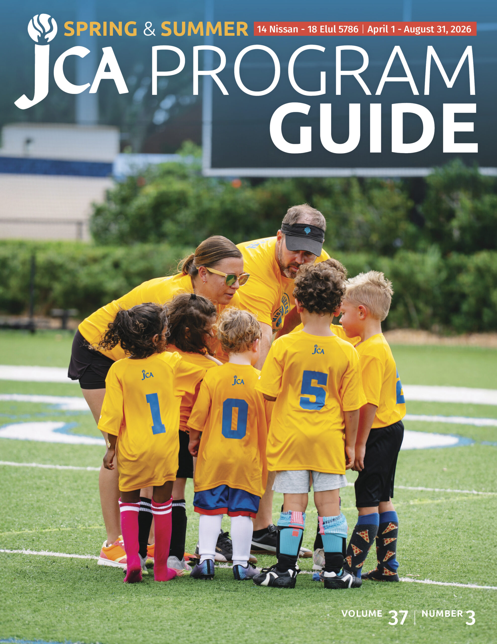A coach kneels and talks to a group of young children wearing yellow JCA shirts on a sports field. The children are huddled closely, listening.