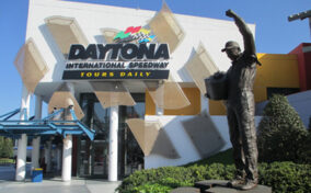 Statue of a race car driver with a raised arm stands in front of the Daytona International Speedway entrance, which has prominent signage and glass features.