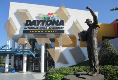 Statue of a race car driver with a raised arm stands in front of the Daytona International Speedway entrance, which has prominent signage and glass features.