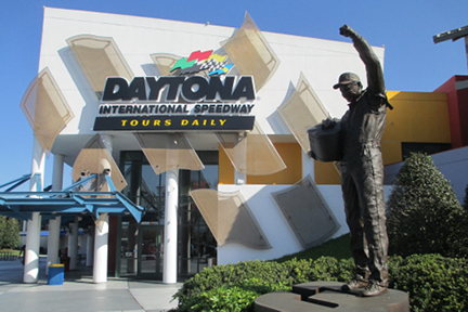 Statue of a race car driver with a raised arm stands in front of the Daytona International Speedway entrance, which has prominent signage and glass features.
