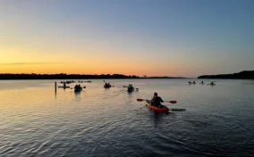 A group of people kayak on calm water at sunset, with the sky glowing orange and blue near the horizon.