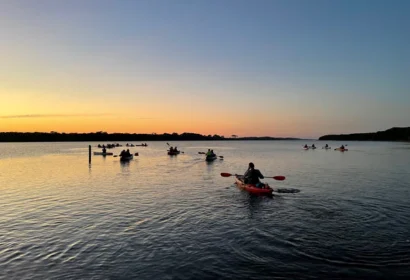 A group of people kayak on calm water at sunset, with the sky glowing orange and blue near the horizon.