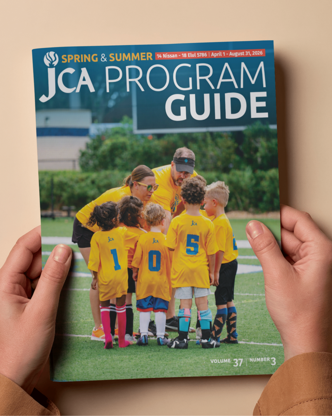 Person holding a JCA Program Guide featuring a group of young children in yellow jerseys with numbers, standing with adult coaches on a grassy field.