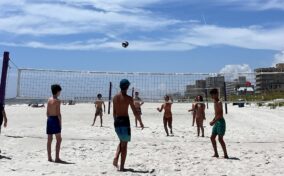 A group of children play beach volleyball on a sandy court under a clear blue sky, with buildings visible in the background.