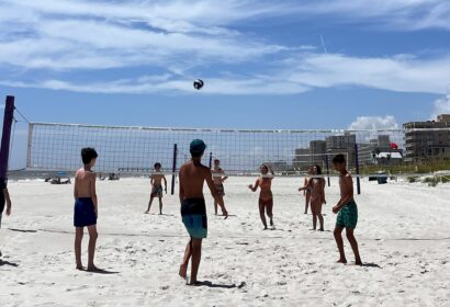 A group of children play beach volleyball on a sandy court under a clear blue sky, with buildings visible in the background.