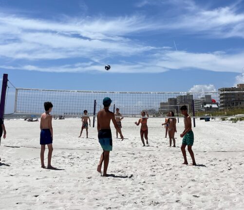 A group of children play beach volleyball on a sandy court under a clear blue sky, with buildings visible in the background.