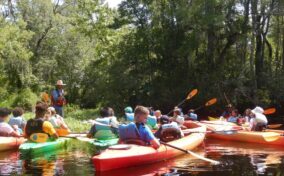 A group of people in colorful kayaks gather on a calm river surrounded by trees, listening to a guide in a straw hat.