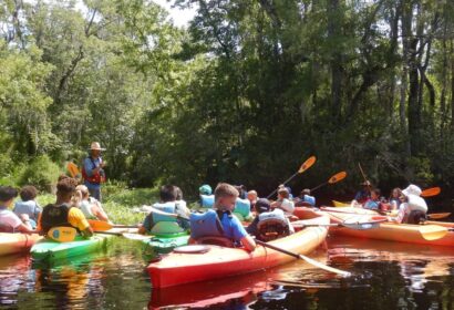 A group of people in colorful kayaks gather on a calm river surrounded by trees, listening to a guide in a straw hat.