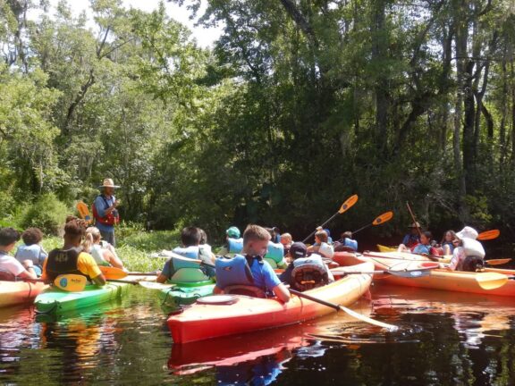 A group of people in colorful kayaks gather on a calm river surrounded by trees, listening to a guide in a straw hat.