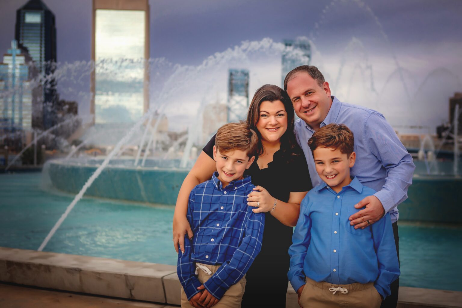 A family of four poses and smiles in front of a large fountain, with tall buildings and a cloudy sky in the background.