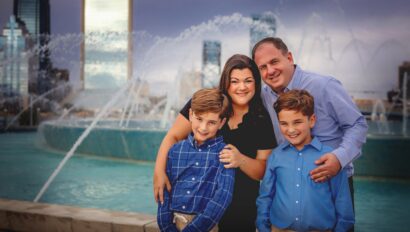 A family of four poses and smiles in front of a large fountain, with tall buildings and a cloudy sky in the background.