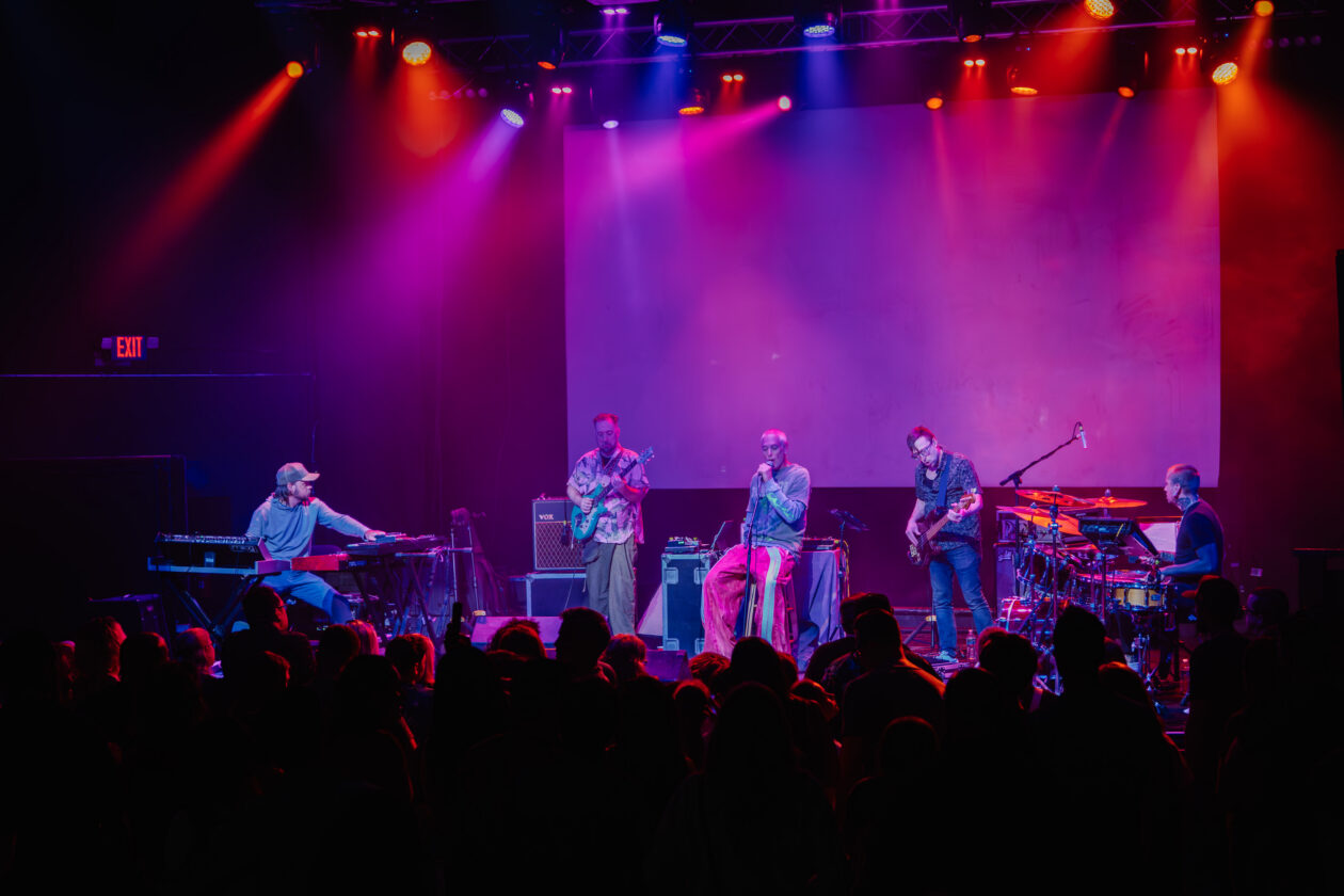 Five musicians perform on stage with instruments including keyboard, guitar, bass, and drums under colorful stage lights, while an audience watches in the foreground.