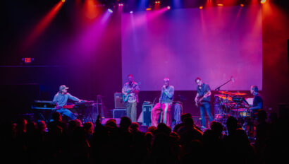 Five musicians perform on stage with instruments including keyboard, guitar, bass, and drums under colorful stage lights, while an audience watches in the foreground.