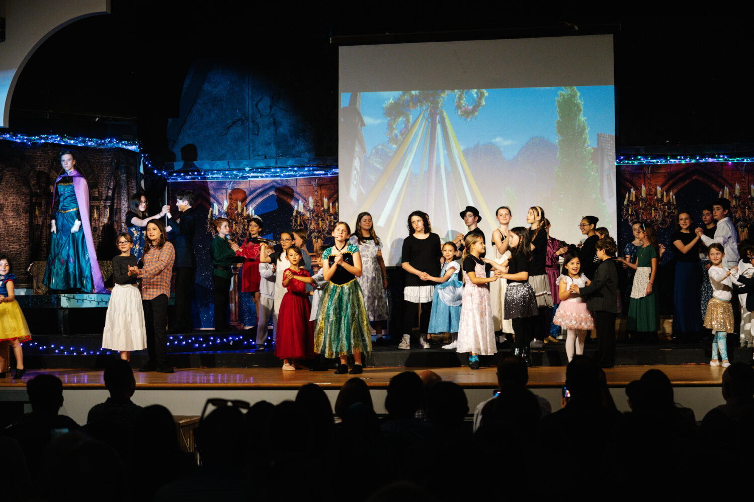 A large group of children and adults in costumes perform on stage, with a colorful backdrop and audience watching in the foreground.