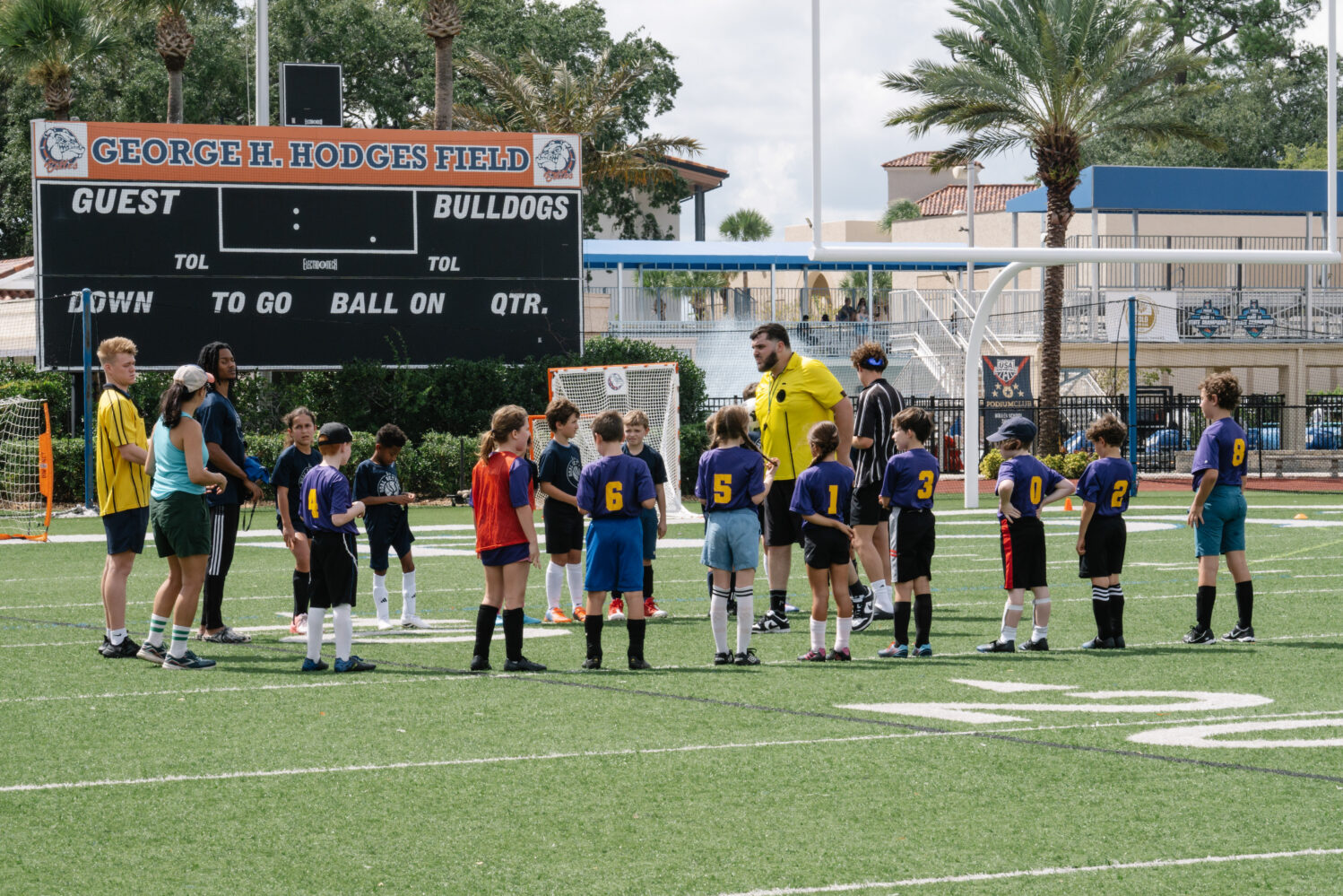 Children in soccer uniforms stand in a circle on a field with coaches and referees, near a scoreboard that reads "George H. Hodges Field.