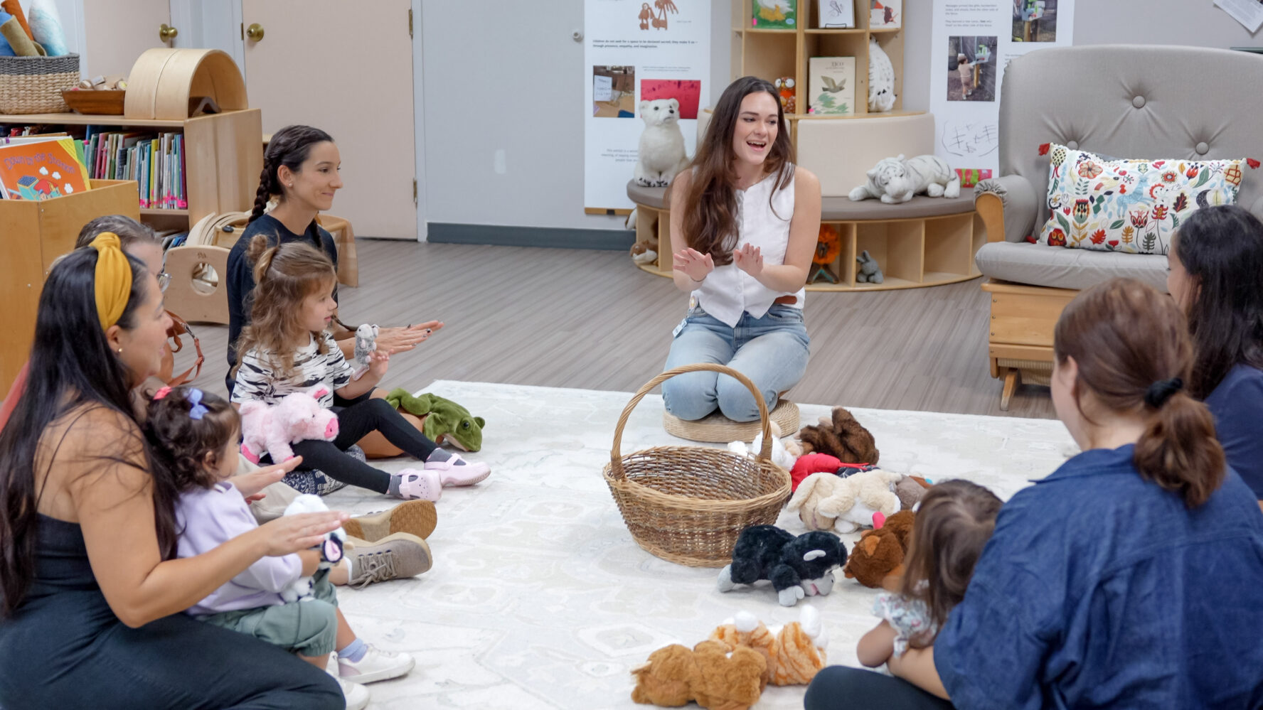 An adult leads a group of young children and their caregivers in a circle activity, surrounded by plush toys and a basket, in a brightly lit classroom.