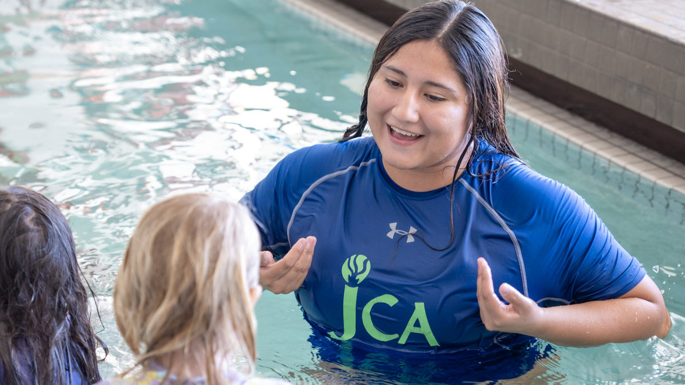 A swim instructor in a blue JCA shirt talks to two children in a swimming pool near the pool’s edge.