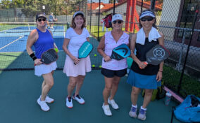 Four women in athletic wear stand on a pickleball court, each holding a paddle and smiling at the camera, with a net and another court visible in the background.