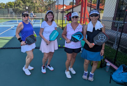 Four women in athletic wear stand on a pickleball court, each holding a paddle and smiling at the camera, with a net and another court visible in the background.