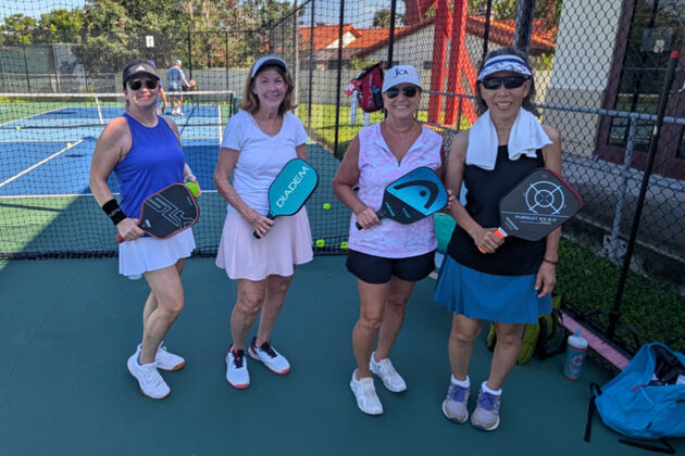 Four women in athletic wear stand on a pickleball court, each holding a paddle and smiling at the camera, with a net and another court visible in the background.