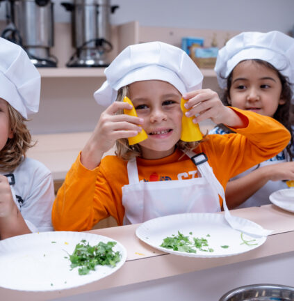 Three children wearing chef hats and aprons sit at a counter with plates of chopped herbs; the child in the center holds two yellow bell pepper slices to their face.