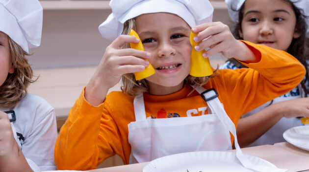Three children wearing chef hats and aprons sit at a counter with plates of chopped herbs; the child in the center holds two yellow bell pepper slices to their face.