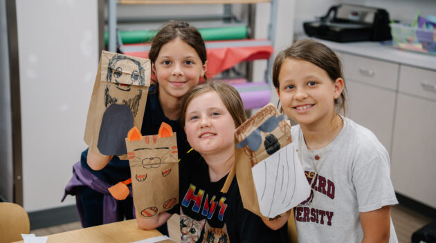 Three children sit at a table holding up paper bag puppets they made, smiling at the camera in a classroom setting.