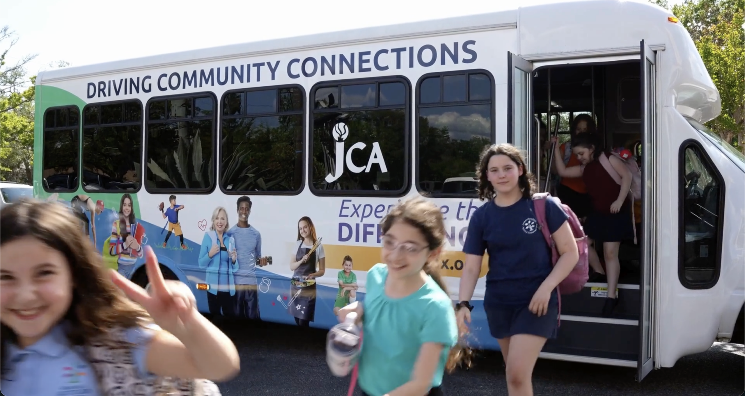 Children exit a JCA community connections bus, with some walking away and others smiling toward the camera.