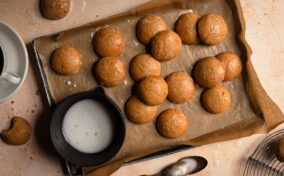 A baking sheet with round cookies on parchment paper, a cup of coffee, a bowl of icing, a bitten cookie, and two spoons on a tan surface.