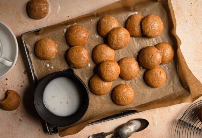 A baking sheet with round cookies on parchment paper, a cup of coffee, a bowl of icing, a bitten cookie, and two spoons on a tan surface.