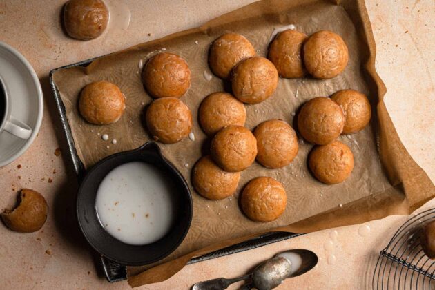 A baking sheet with round cookies on parchment paper, a cup of coffee, a bowl of icing, a bitten cookie, and two spoons on a tan surface.