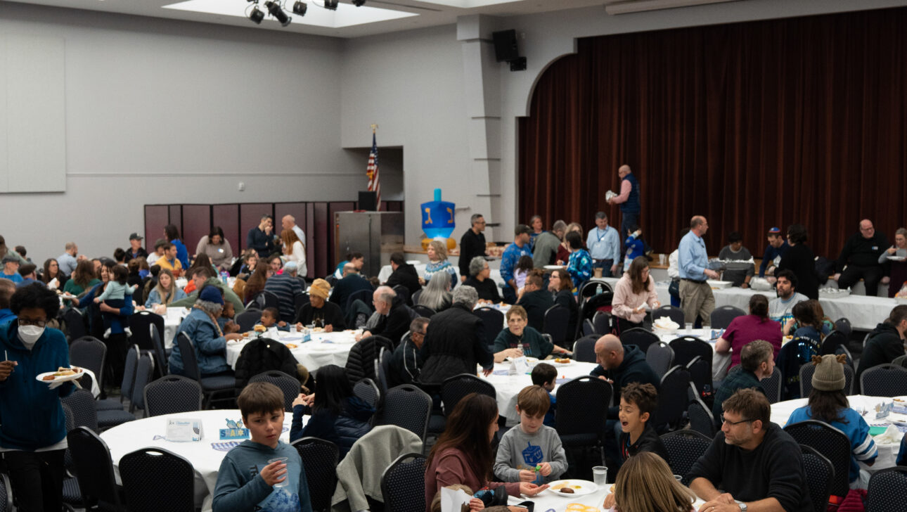 A large group of people sit at round tables in a spacious hall, eating and talking. Some are standing or walking around. There is a stage with a closed curtain in the background.
