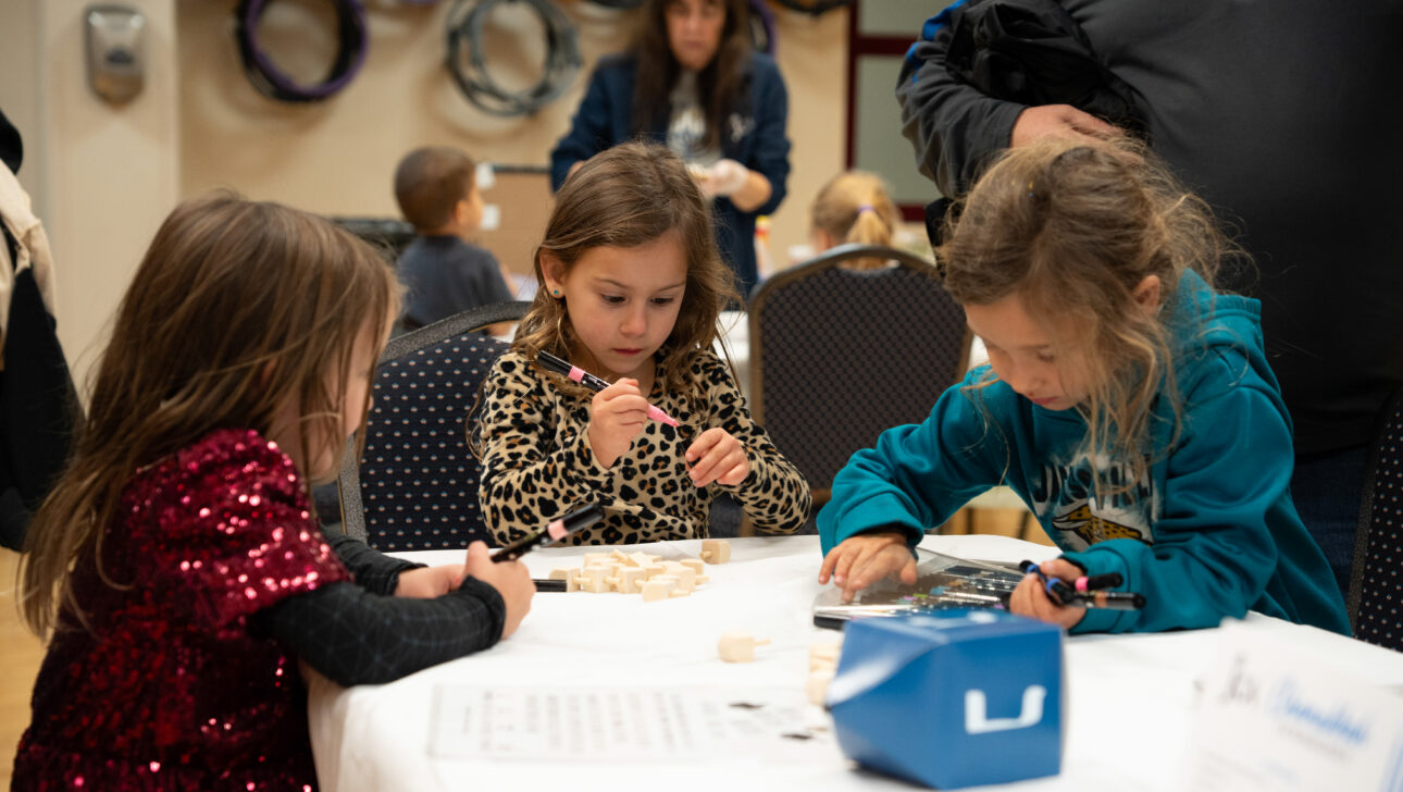 Three young children sit at a table working on crafts with wooden pieces and markers. Other children and adults are visible in the background.
