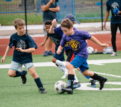 Four boys in sportswear compete for a soccer ball on a green field. One boy in a purple shirt leads, while others in blue shirts follow closely. Fence and running track visible in background.
